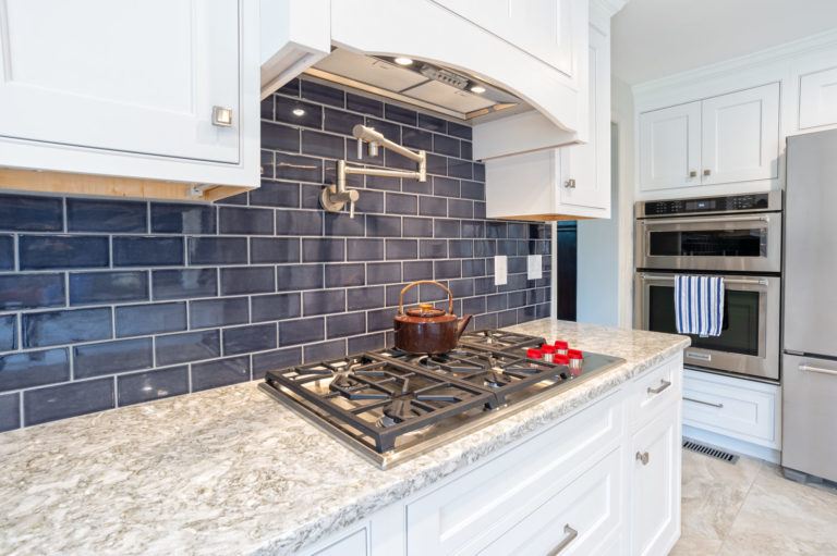 Dark blue backsplash behind a gas range a pot filler and white vent hood built into the cabinetry.